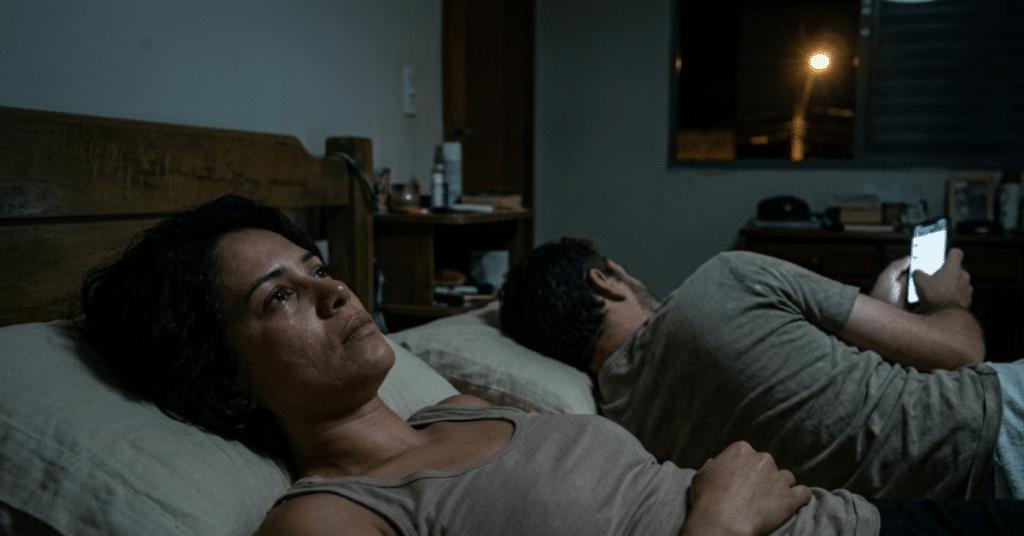 Fadiga emocional no casamento: mulher acordada e chorando na cama enquanto o marido está de costas mexendo no celular no escuro, ilustrando a distância de colegas de quarto.